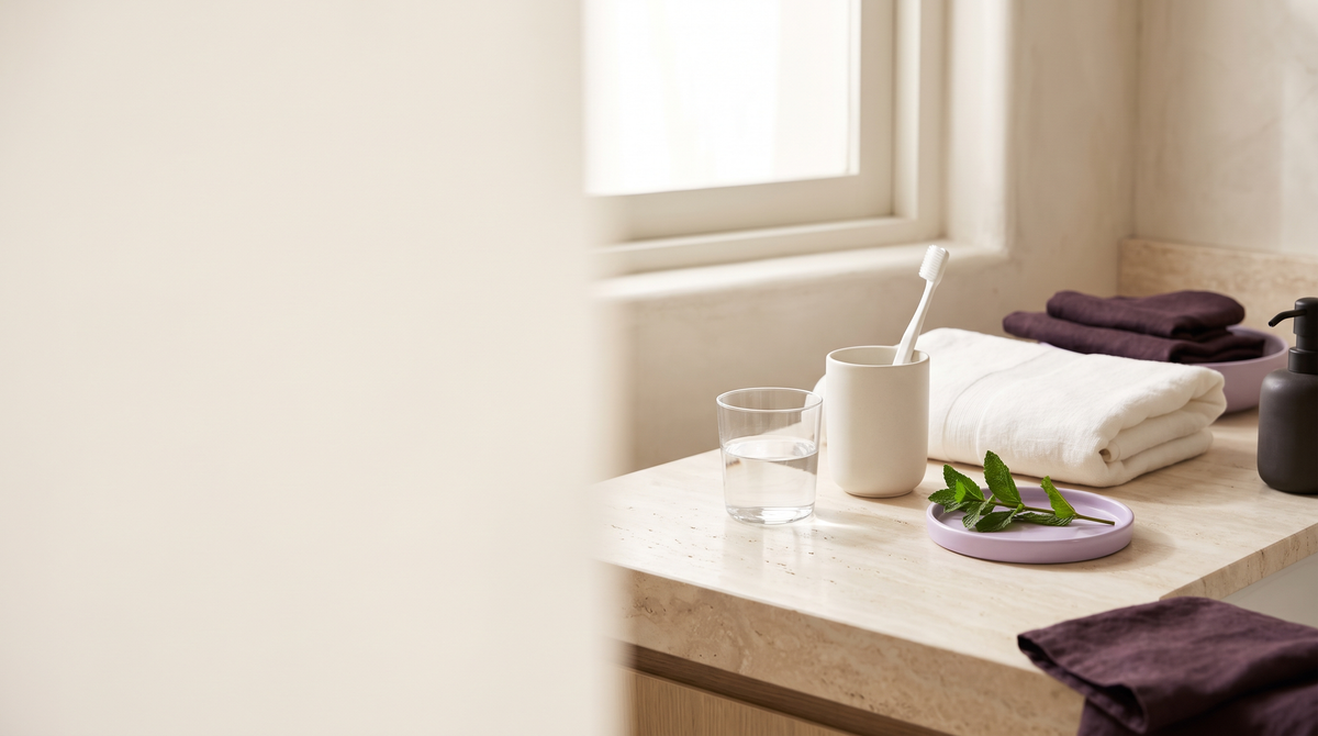 A calm bathroom vanity in morning light with a glass of water, a toothbrush, folded towels and fresh mint