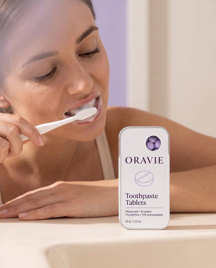 Step 2, brush: woman brushing her teeth with a white toothbrush, ORAVIE tin on the counter
