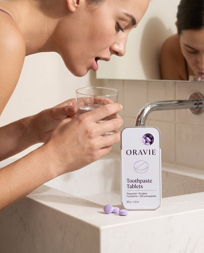 Step 3, rinse: woman rinsing with a glass of water at a stone sink, ORAVIE tin on the edge